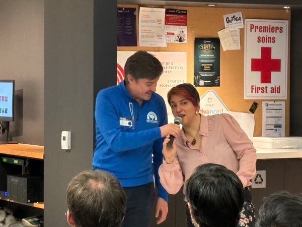 A candid indoor photograph captures a woman speaking into a microphone next to a man wearing a blue branded shirt, standing in front of a cork bulletin board during an event. The foreground shows the backs of several audience members' heads.