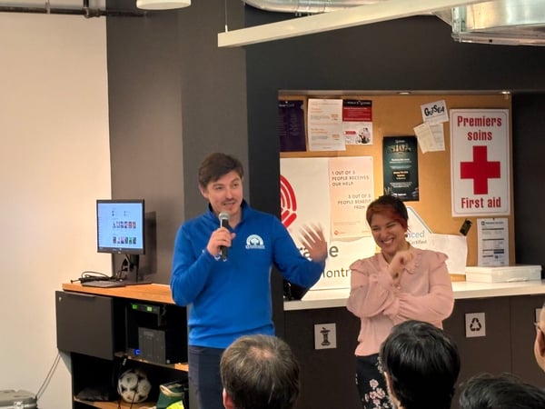 A candid photo taken indoors showing a man in a blue shirt speaking into a microphone during a presentation, while a woman beside him laughs enthusiastically. They stand in front of a dark wall featuring a monitor displaying graphics and a corkboard with various posters and a bilingual First Aid sign.