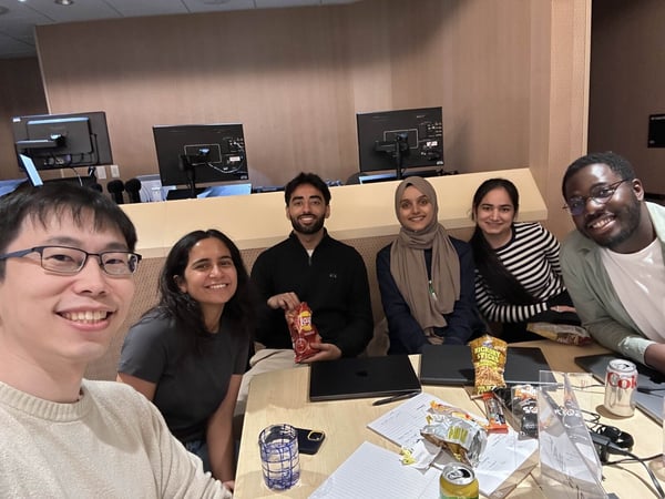 A group selfie features six smiling individuals (three men and three women) gathered around a light wooden table indoors, likely in an office or classroom setting.
