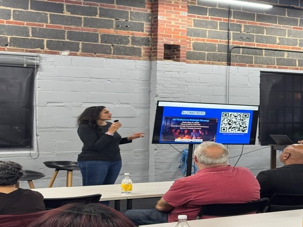 A woman is giving a presentation at an AI Tinkerers Raleigh Meetup, standing in front of a screen displaying event details and a QR code.
