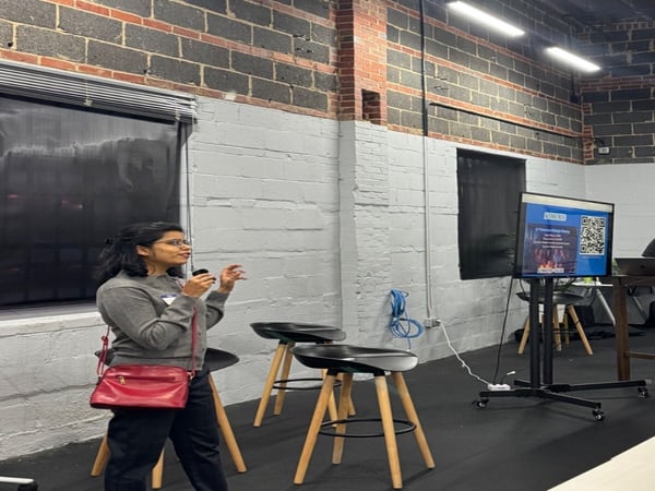 A woman is giving a presentation in an indoor space with a brick wall, standing near a monitor displaying a QR code and several stools.