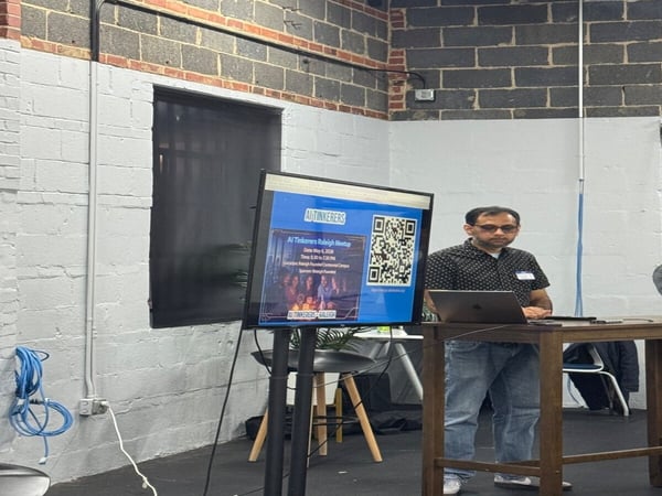 A man sits at a wooden table with a laptop, positioned in front of a monitor displaying a slide for an 'AI Tinkerers Raleigh Meetup'. The setting is an indoor space with white-painted and exposed brick walls.