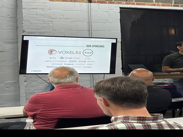A photo of an indoor presentation where several people are seated, looking at a screen displaying a list of sponsors like VOXEL51 and Google Cloud. The image is oriented sideways.