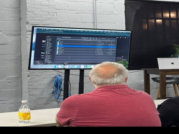 A man in a red shirt is seen from behind, sitting at a desk and looking at a large computer monitor displaying a macOS file interface. The setting is an indoor space with a white-painted brick wall.