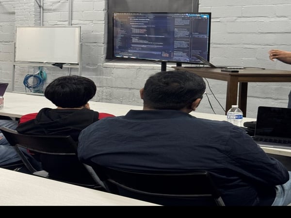 A group of people are seated at a table in a room with brick walls, focused on a large monitor displaying computer code.