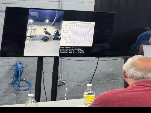 A sideways photograph showing a person watching a monitor that displays a weightlifting video alongside performance metrics and a displacement graph.