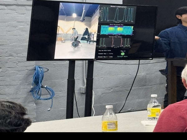 A photograph showing a person giving a demonstration or presentation using a large monitor that displays video of a person weightlifting along with various data visualizations like graphs and heatmaps.