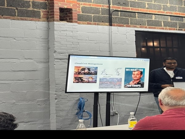 The image shows a man giving a presentation in a room with a brick wall. A monitor displays a slide titled 'Cloud Cost Management' featuring several memes.