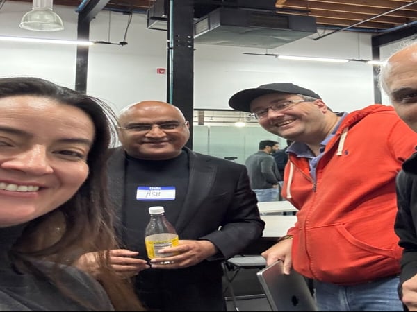 A group selfie of four people smiling in an indoor setting with high ceilings and exposed wooden beams. One person wears a name tag that says 'ASH' and another holds a tablet.