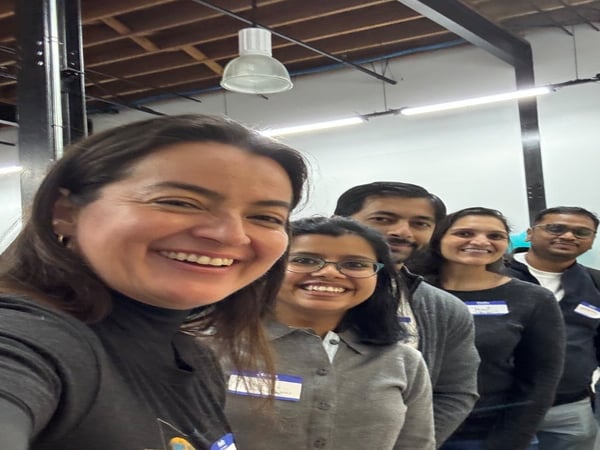 A group of five smiling people, three women and two men, posing for a selfie in an indoor setting with high ceilings and exposed beams.