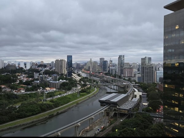 A wide-angle photograph capturing a dense urban landscape under a heavily overcast sky, featuring a river, elevated train tracks leading to a station, and numerous high-rise buildings, including a large, dark glass skyscraper dominating the right foreground.
