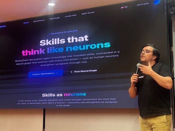 A man is giving a presentation in front of a large screen displaying a website for 'SkillsChain' with the headline 'Skills that think like neurons'.
