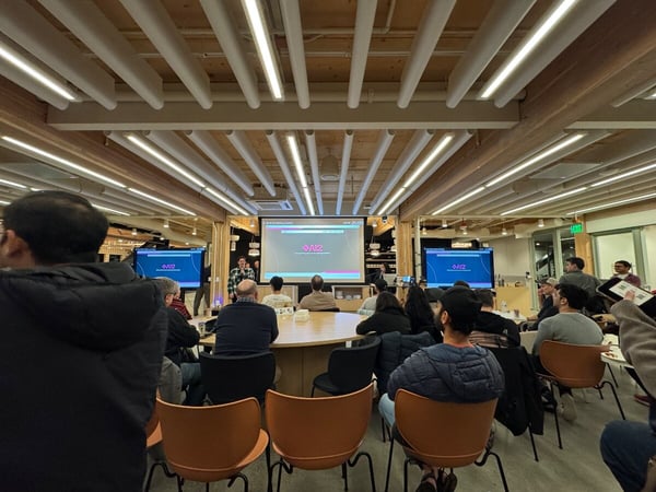 A candid photograph captures an indoor presentation or meeting where a large audience is seated around a table, facing a central projection screen and two side monitors, all displaying the AI2 logo.