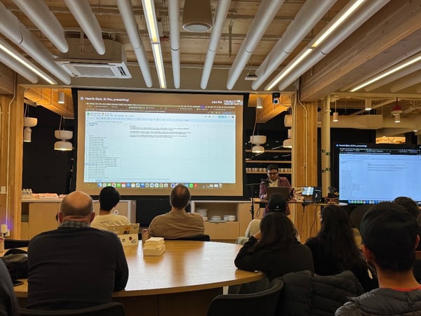 A photograph capturing an indoor presentation or technical meeting where attendees are watching a presenter display a spreadsheet or data list on a large projection screen.