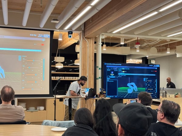 A photograph captures an indoor technical presentation where a presenter stands at a table demonstrating robotics software displayed on two large screens to an audience seated in the foreground. The setting features modern wooden architecture and exposed ceilings.