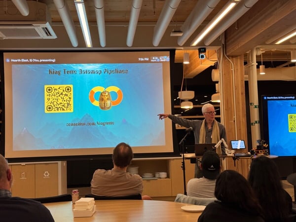 A man is giving a presentation titled "King Tut Datamap Pipelines" in a modern indoor venue, pointing toward the large projection screen that displays a QR code and a graphic of the Tutankhamun mask. Several audience members are seated in the foreground, watching the presentation.