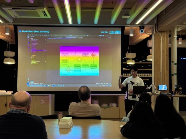A photograph captures an indoor technical presentation where a speaker, standing next to a large projection screen displaying a colorful flowchart and code, addresses a small audience seated at a wooden table.