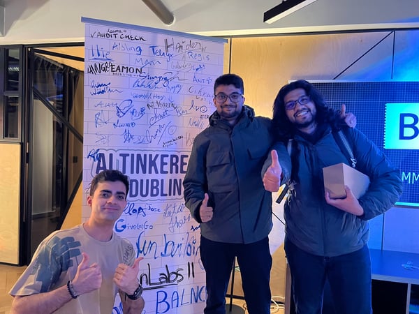 Three young men pose together smiling and giving thumbs-up gestures next to a large banner covered in handwritten signatures and names.