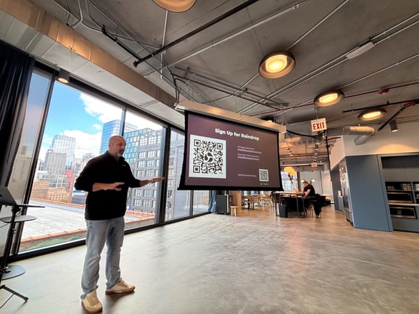 A man in casual attire is giving a presentation in a modern office space with large windows overlooking a city skyline, standing next to a projection screen displaying text and QR codes related to signing up for Raindrop and Chicago Data Portal resources.