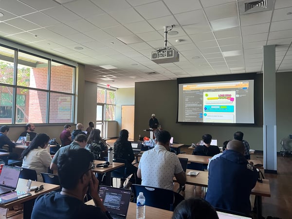 A group of students or participants are seated at desks in a classroom-style setting, focused on a presentation being shown on a large projection screen at the front of the room.