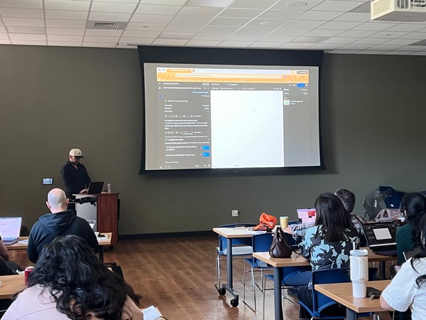 A man in a baseball cap gives a presentation to an audience in a room with a large projector screen displaying a GitHub pull request.