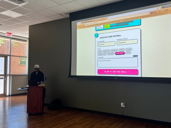 A person in a baseball cap stands at a podium giving a presentation, with a large projector screen behind them displaying a hackathon guestbook web application.