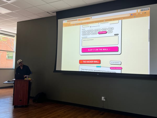 A man stands at a podium giving a presentation in a room, with a large screen behind him displaying a web application interface.