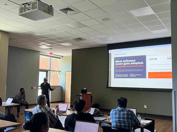 A man stands at the front of a room giving a presentation to an audience, with a large projector screen displaying hackathon information.