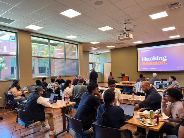 A group of people are gathered in a brightly lit room for a 'Hacking Session,' working on laptops at several tables while a projector displays the event details.