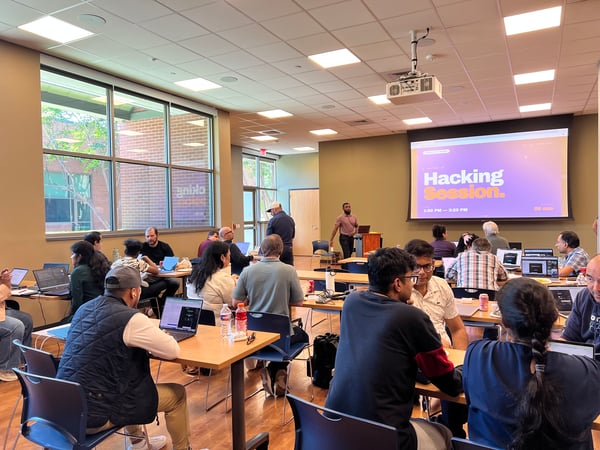 A group of people are gathered in a large, well-lit room for a 'Hacking Session' workshop, with many participants working on laptops at tables.