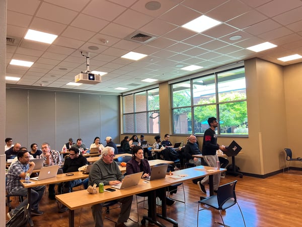 A group of people are seated at long wooden tables in a modern classroom or workshop setting, most of them working on laptops. One person is standing and walking across the room carrying a laptop.