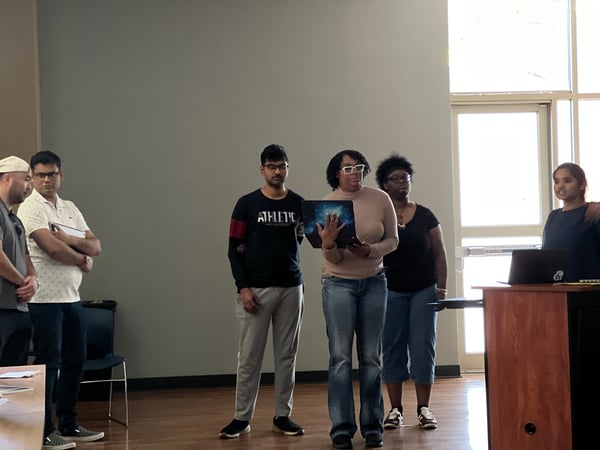 A group of people are gathered in a room for what appears to be a presentation, with one woman speaking from a podium and another holding a laptop.