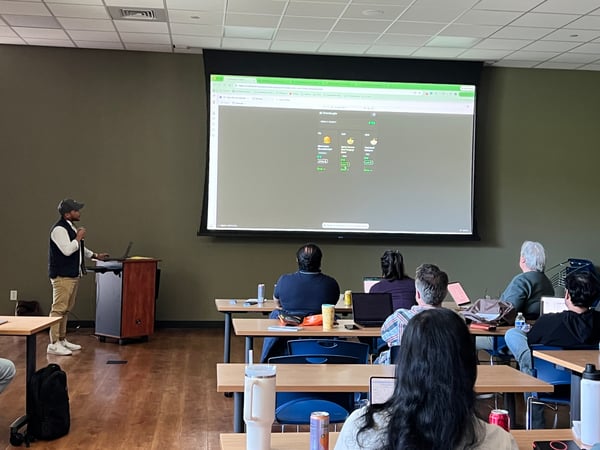 A man stands at a podium giving a presentation to an audience in a room with a large screen displaying a food ordering application.