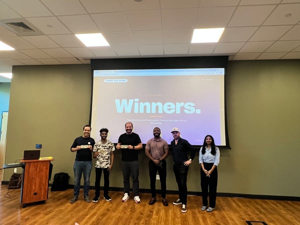 A group of six people posing for a photo in front of a projection screen displaying 'Winners.' in an indoor setting.