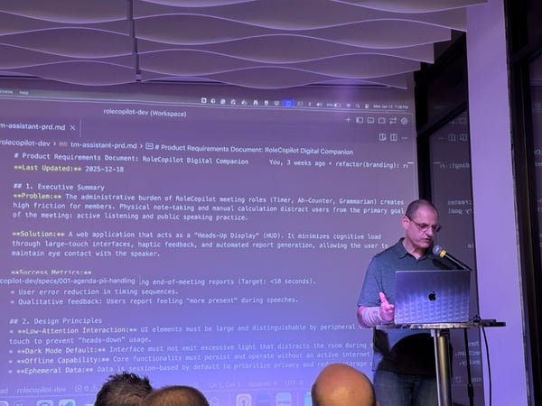 A man is giving a technical presentation in a dimly lit room, standing next to a laptop and microphone while a large screen displays a Product Requirements Document (PRD) with dark mode text.