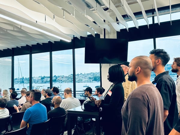 A photograph capturing a large group of people attending an indoor event or presentation, with attendees seated and standing in a modern room featuring large windows overlooking a waterfront cityscape.