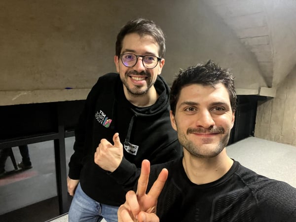 A close-up selfie shows two smiling men indoors, one giving a peace sign and the other a thumbs up, set against a dark, concrete background. The text on the hoodie of the man in the background is reversed due to the camera angle.