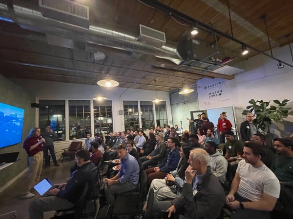 A photograph captures an indoor professional gathering or presentation event taking place at night, showing a woman speaking into a microphone near a large screen while a large audience is seated and listening.
