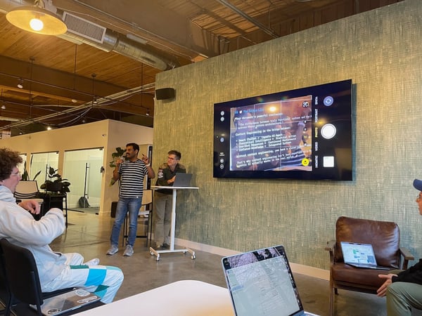 A group of people attends an indoor presentation in a modern office space, where a speaker addresses the audience next to a large wall-mounted screen displaying a slide that is being viewed through a smartphone camera interface.