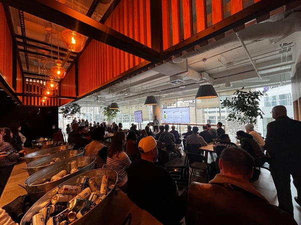 A large group of people gathered in a modern indoor space for a presentation, with a speaker standing before digital screens and drinks available in the foreground.