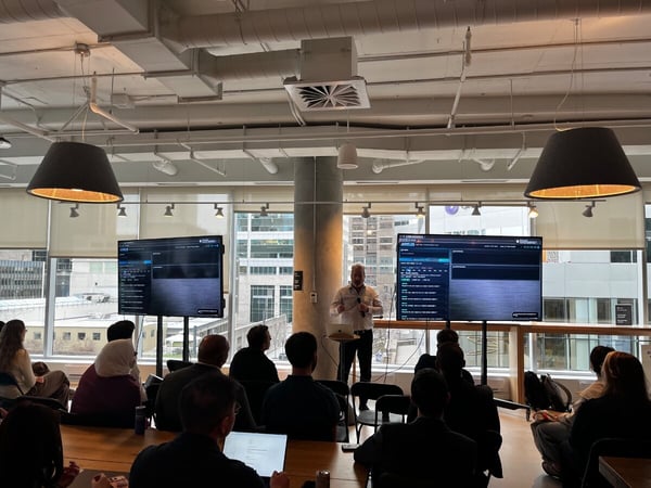 A man is giving a presentation in a modern office space with two large screens displaying software interfaces to a seated audience.