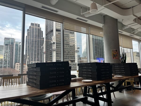 Stacks of black pizza boxes labeled 'SLICE + SODA' sit on a long wooden table in a modern office or restaurant with a view of city skyscrapers.