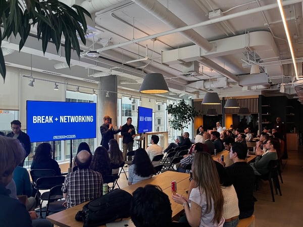 A wide shot of a modern office space where a group of people are attending an event, with presenters standing next to a screen announcing a networking break.