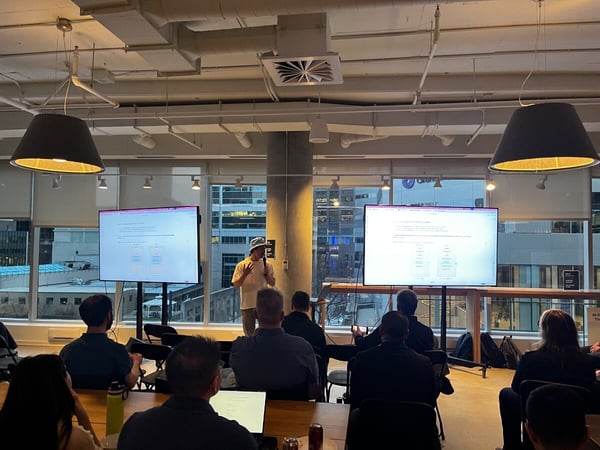 A man in a bucket hat delivers a presentation to an audience in a modern office space with large windows. Two large monitors display technical diagrams and text related to AI and Transformer architectures.