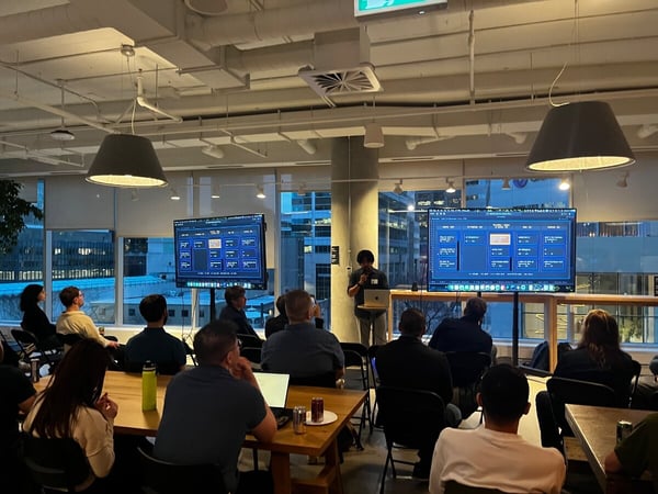 A man gives a presentation in a modern office space to a seated audience, with two large screens displaying project management boards.