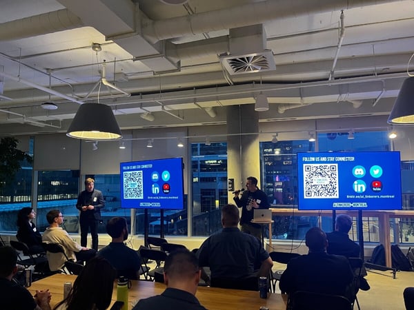 A man is giving a presentation to an audience in a modern office space at night, with large windows showing a city view and two screens displaying social media links.
