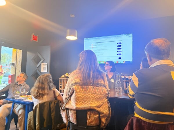 A group of people are gathered in an indoor space, likely a restaurant or event venue, watching a presentation displayed on a large screen.