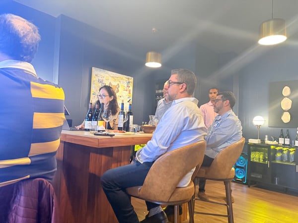 A group of people are gathered around a wooden bar table, likely for a wine tasting or social event, with several wine bottles and glasses visible.