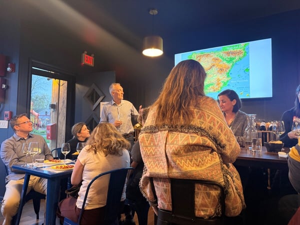 A group of people are gathered in a restaurant or bar for a presentation or social event, with a man standing and speaking while a map of Spain is displayed on a screen.
