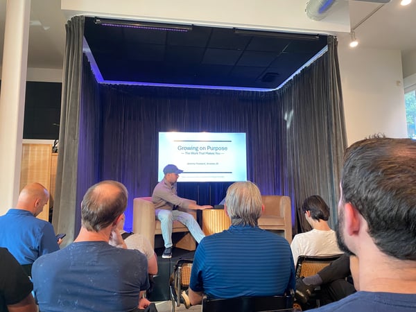 A photograph showing an indoor presentation event where a speaker is seated on a small stage in front of a screen displaying a presentation titled 'Growing on Purpose'. An audience is seated in the foreground with their backs to the camera, watching the speaker.